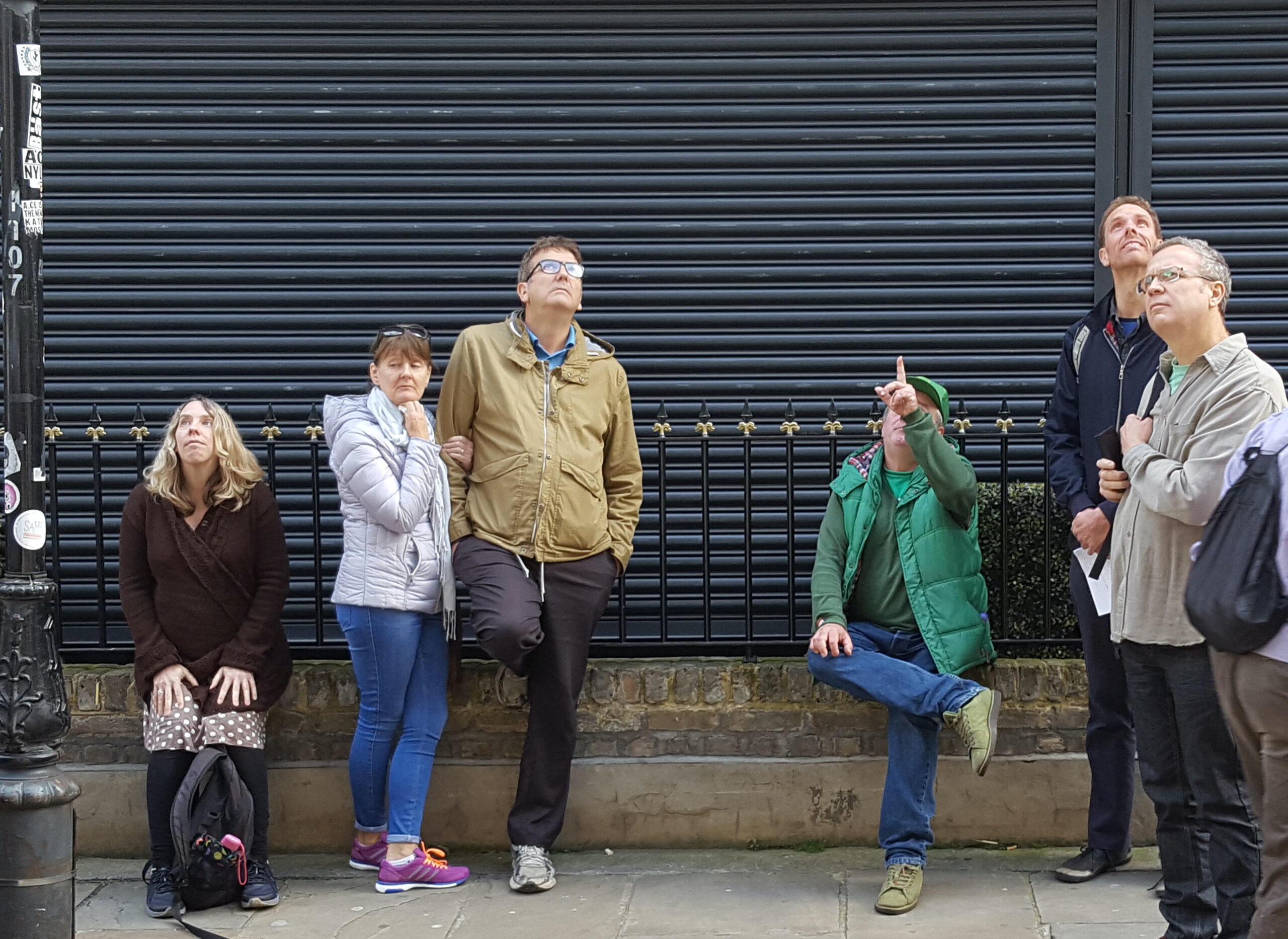 Five people listening to an Unseen Tours guide, Pete on Brick Lane. Pete points upwards to a building as he explains its history to his guests.