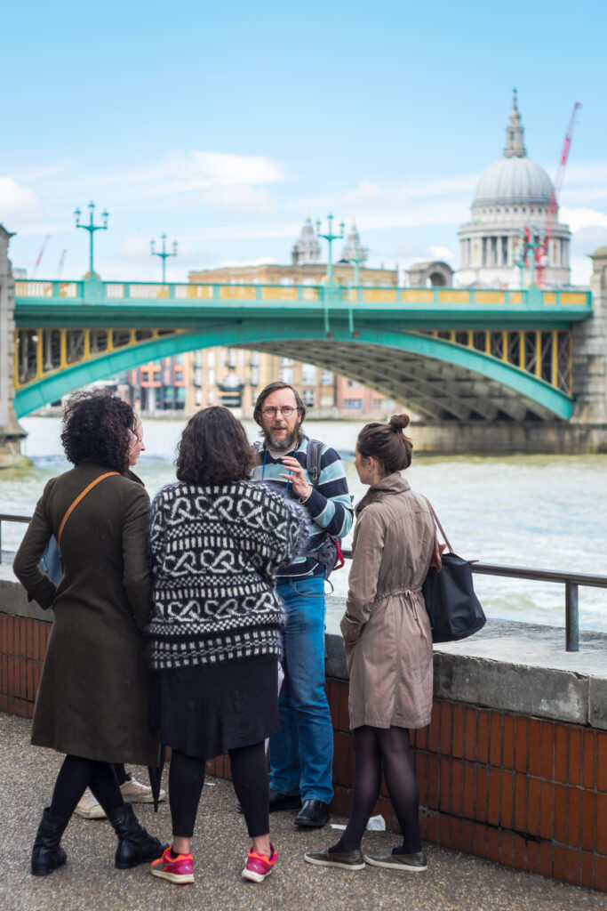 David stands facing three guests as they stand along the Thames River on his tour