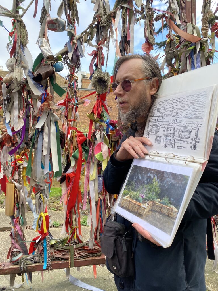 David stands outside of Crossbones Graveyard, holding open his guide book with a photo of the bench inside the graveyard.
